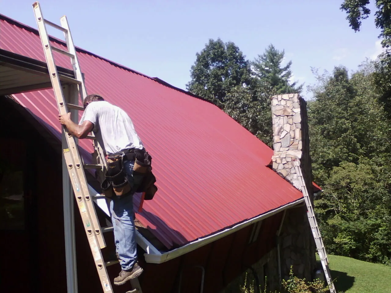 Roofer on a ladder beside a red metal roof and stone chimney — Soffit & Fascia Repair by Roofing Brothers in Mullins